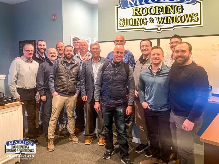 The TrussPoint team poses with the Marios Roofing, Siding & Windows team inside the Marios office during a partner visit, with the company's branded sign visible on the wall behind the group.