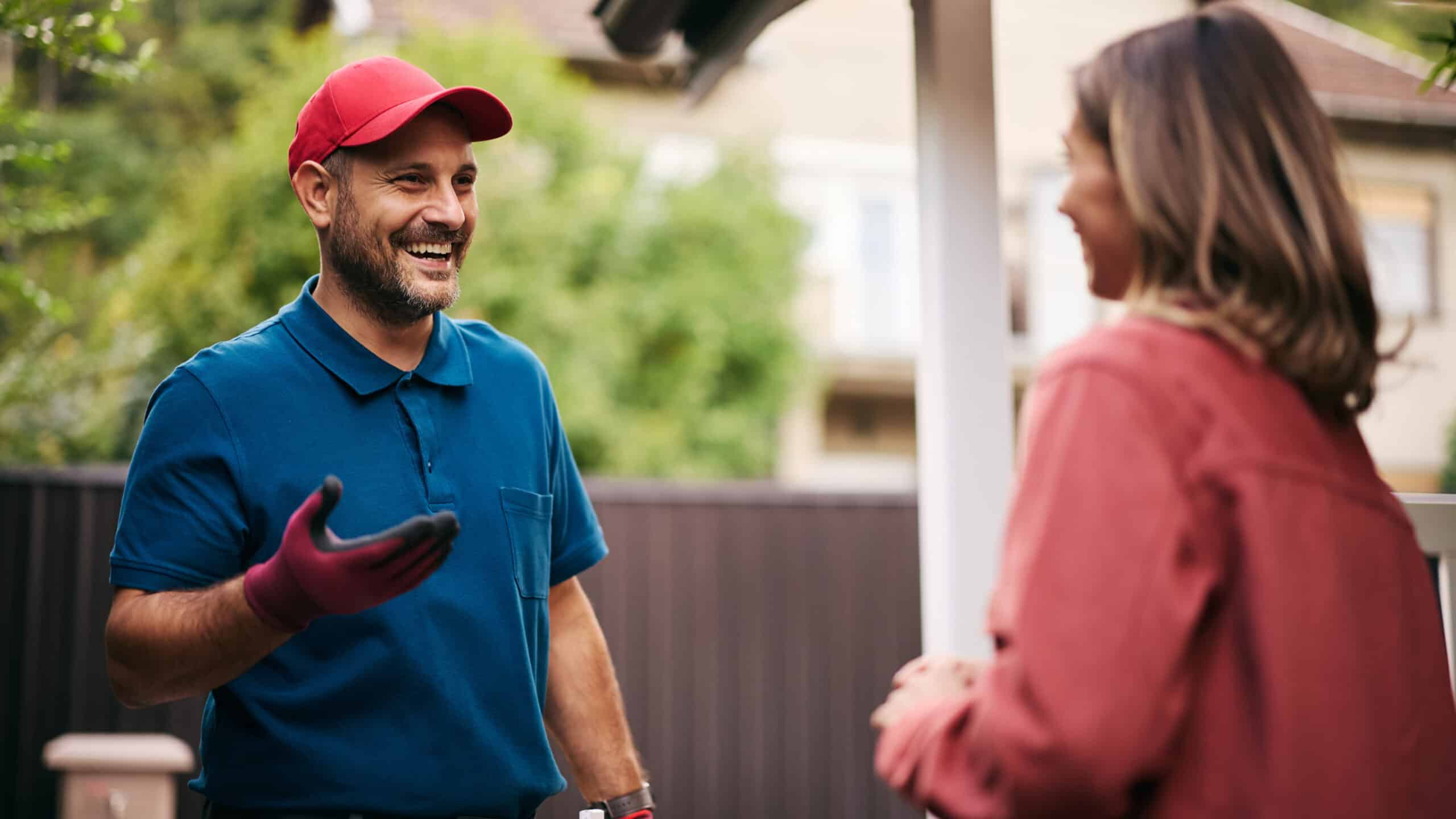 Smiling contractor in a red cap, blue polo, and work gloves chatting with a homeowner outside her house.