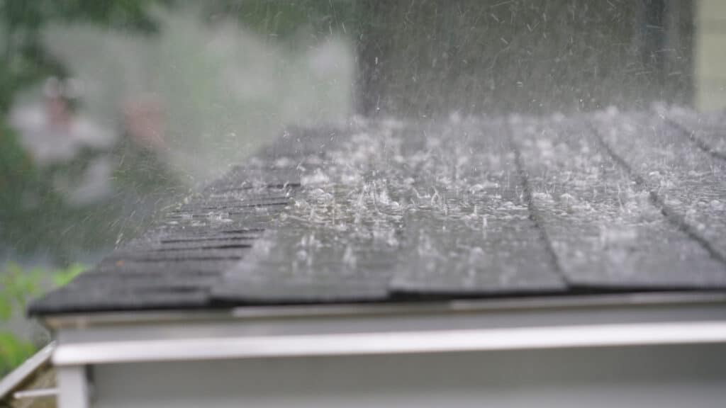 Close-up of heavy rain pounding a dark asphalt shingle roof, with water splashing up from the shingles above the gutter line.