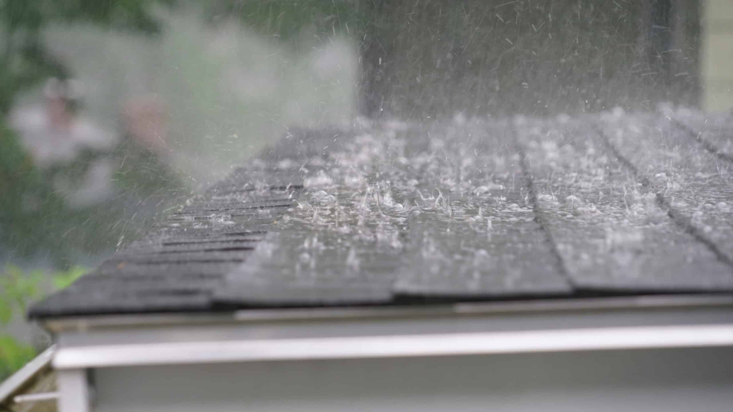 Close-up of heavy rain pounding a dark asphalt shingle roof, with water splashing up from the shingles above the gutter line.