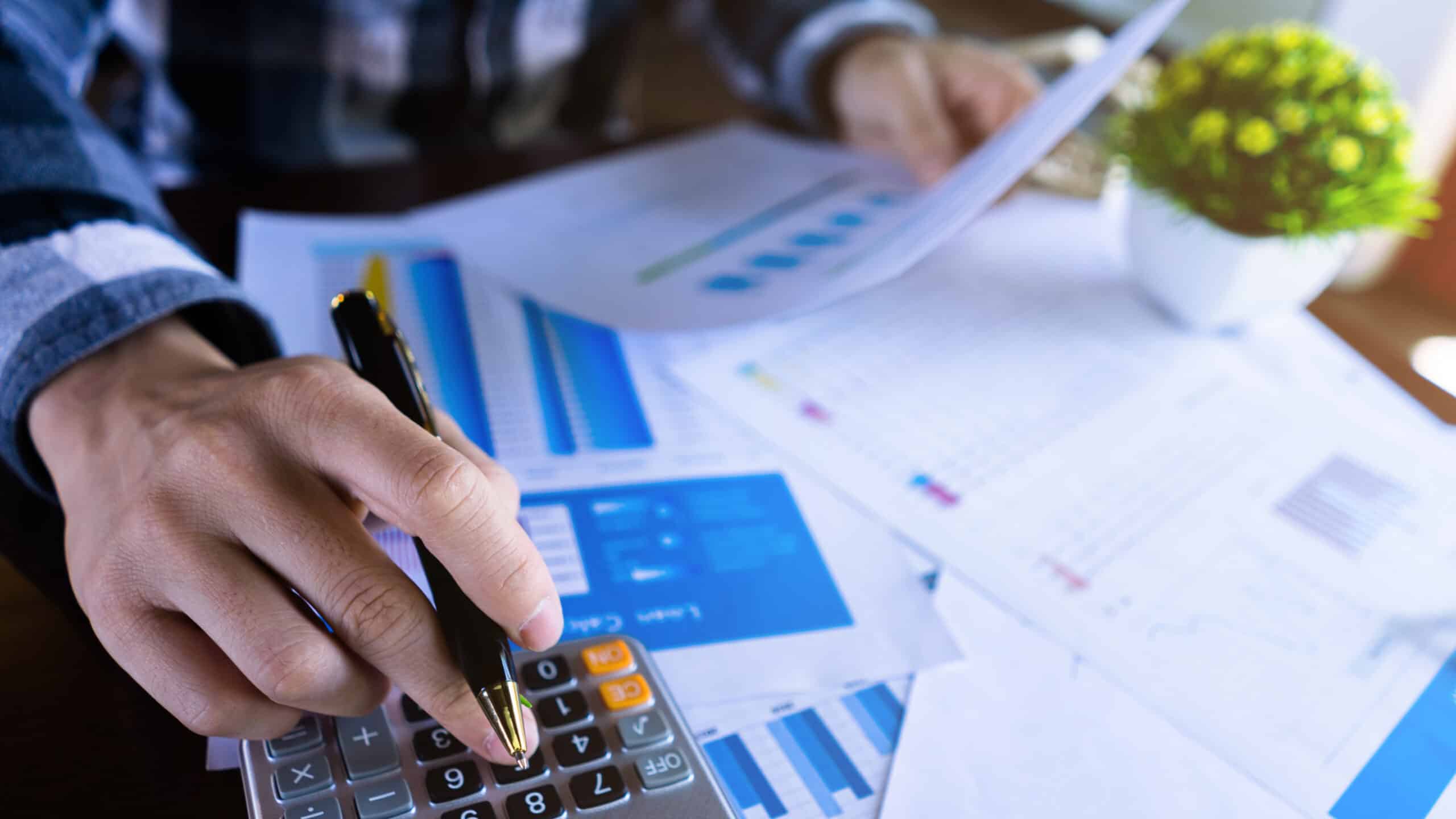 Close-up of a business owner using a pen to tap a calculator while reviewing printed financial reports with bar charts and spreadsheets.